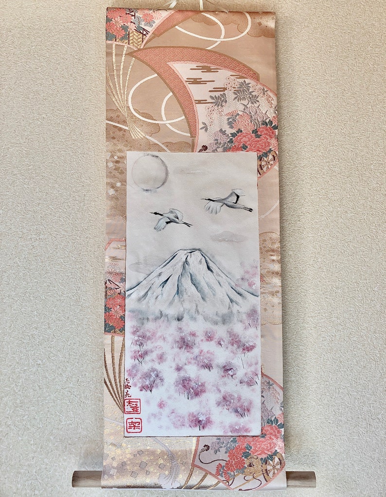 May include: Japanese wall scroll depicting Mount Fuji, cherry blossoms, cranes, and a full moon. The scroll has a beige and pink patterned fabric border with floral and fan motifs. A wooden dowel is at the bottom.