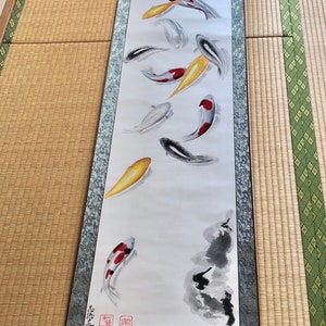 May include: A long, vertical scroll painting featuring koi fish in shades of red, yellow, black, and white. The artwork is mounted on a blue-gray fabric with a wooden roller at the bottom. The background is white, and the style is reminiscent of traditional Japanese art.