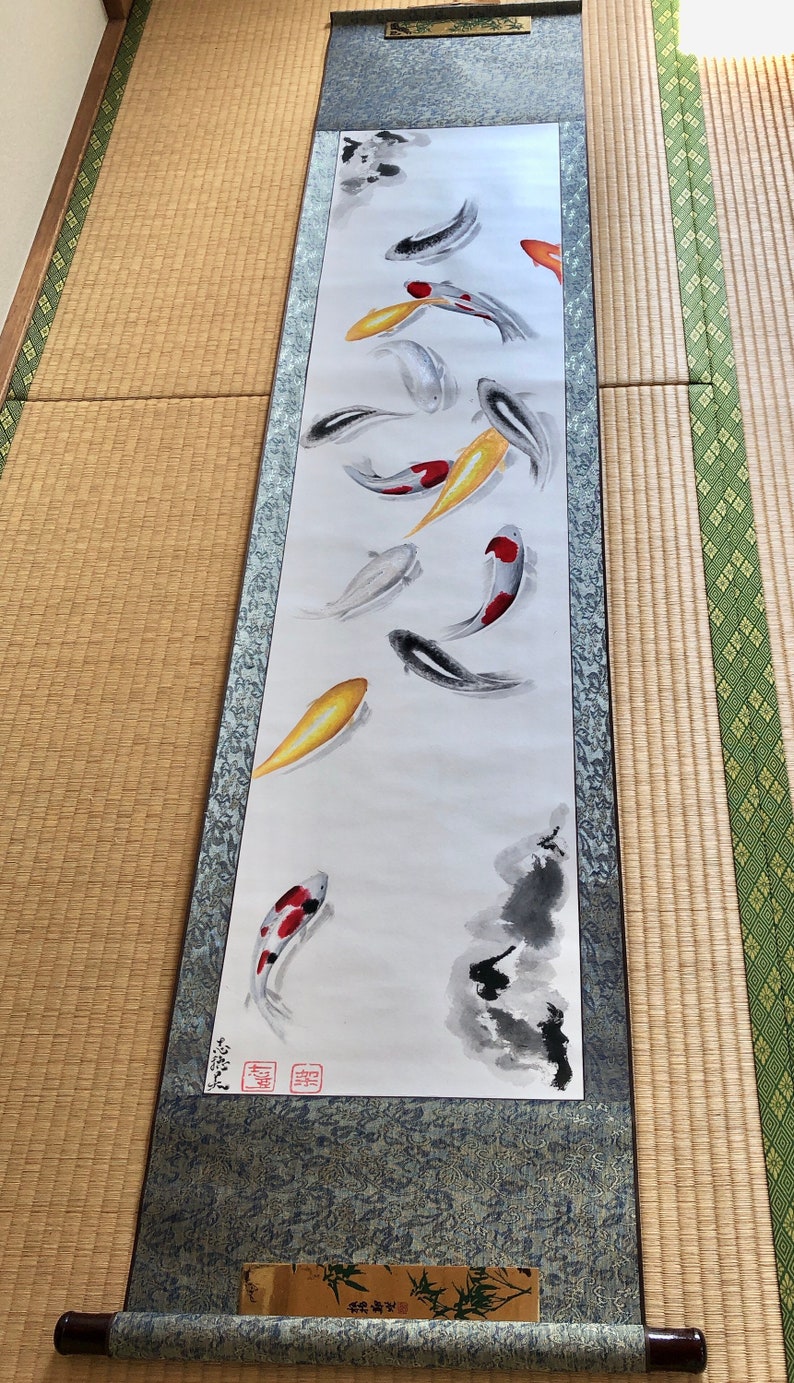 May include: A long, vertical scroll painting featuring koi fish in shades of red, yellow, black, and white. The artwork is mounted on a blue-gray fabric with a wooden roller at the bottom. The background is white, and the style is reminiscent of traditional Japanese art.
