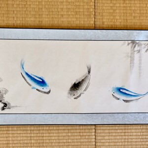 May include: A long, rectangular watercolor painting featuring three fish in shades of blue and gray. The fish are swimming in a light background with bamboo stalks on the right. The painting has a light blue border and is framed.