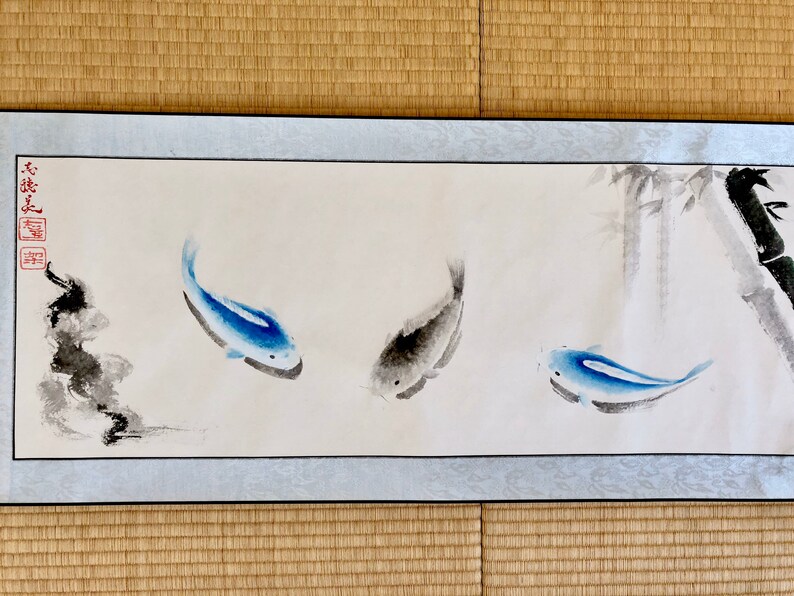 May include: A long, rectangular watercolor painting featuring three fish in shades of blue and gray. The fish are swimming in a light background with bamboo stalks on the right. The painting has a light blue border and is framed.