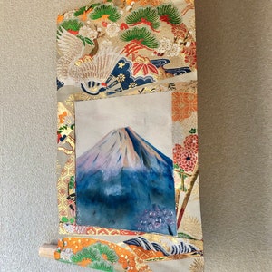 May include: A Japanese scroll painting featuring Mount Fuji. The artwork is framed by ornate fabric with gold accents, floral patterns, and a crane design. The scroll is suspended by a gold ribbon and has a wooden dowel at the bottom.