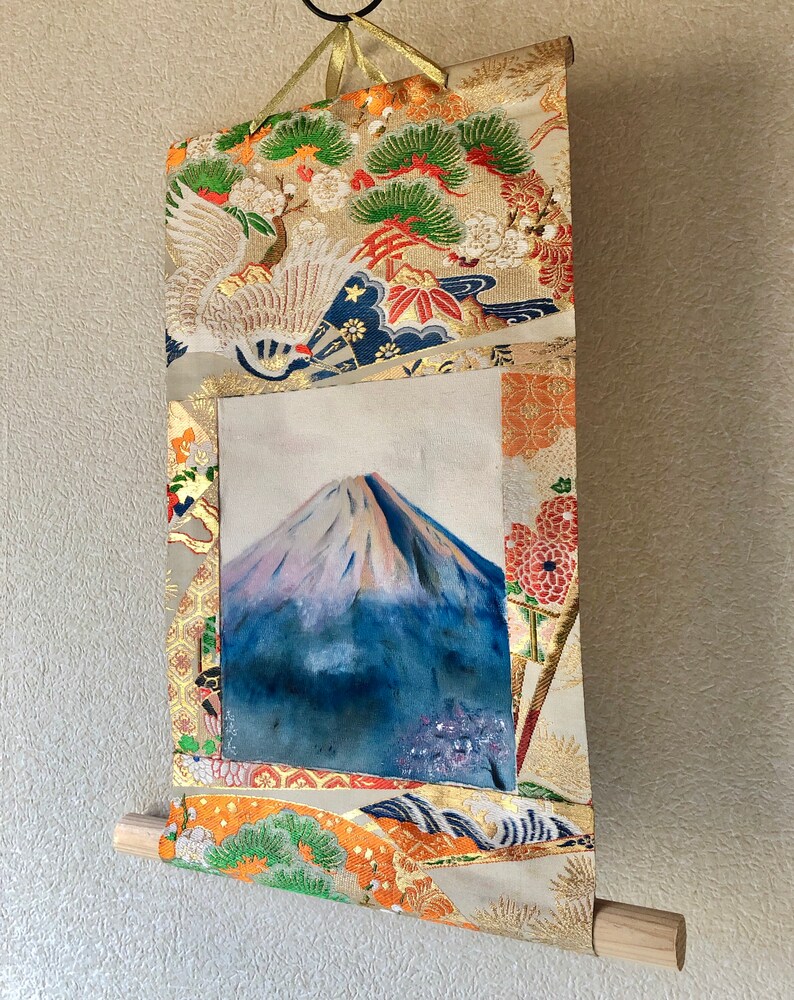 May include: A Japanese scroll painting featuring Mount Fuji. The artwork is framed by ornate fabric with gold accents, floral patterns, and a crane design. The scroll is suspended by a gold ribbon and has a wooden dowel at the bottom.