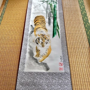 May include: A traditional Japanese scroll painting featuring a tiger with orange and black stripes, walking towards the viewer. The artwork includes bamboo stalks and Japanese characters. The scroll has a gray patterned border and wooden hanging hardware.