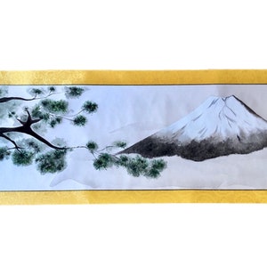 May include: A Japanese scroll painting featuring Mount Fuji with a snow-capped peak and a tree with green foliage. The artwork is framed by a gold border and decorative end pieces. The painting is a traditional landscape scene.