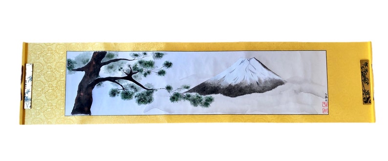 May include: A Japanese scroll painting featuring Mount Fuji with a snow-capped peak and a tree with green foliage. The artwork is framed by a gold border and decorative end pieces. The painting is a traditional landscape scene.