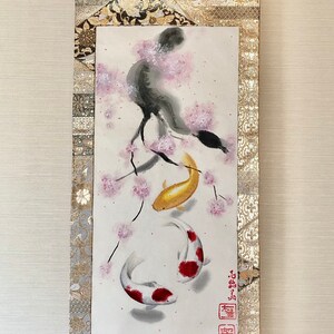 May include: Japanese-style wall scroll featuring a watercolor painting of two koi fish swimming beneath a cherry blossom branch. The artwork is framed by gold and patterned fabric, with a wooden dowel at the bottom.