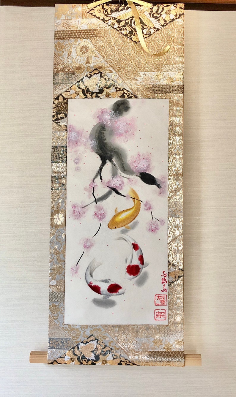 May include: Japanese-style wall scroll featuring a watercolor painting of two koi fish swimming beneath a cherry blossom branch. The artwork is framed by gold and patterned fabric, with a wooden dowel at the bottom.