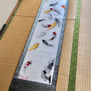 May include: A long, rectangular scroll featuring a watercolor painting of koi fish in shades of red, yellow, black, and white. The scroll has a dark blue patterned border and wooden dowels at the top and bottom.