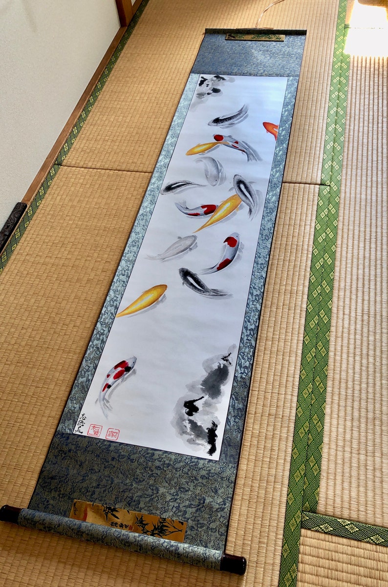 May include: A long, rectangular scroll featuring a watercolor painting of koi fish in shades of red, yellow, black, and white. The scroll has a dark blue patterned border and wooden dowels at the top and bottom.