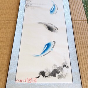 May include: A traditional Japanese scroll painting featuring three koi fish in shades of blue and gray, swimming in a minimalist style. Bamboo stalks are painted at the top. The scroll is framed with a light blue border.