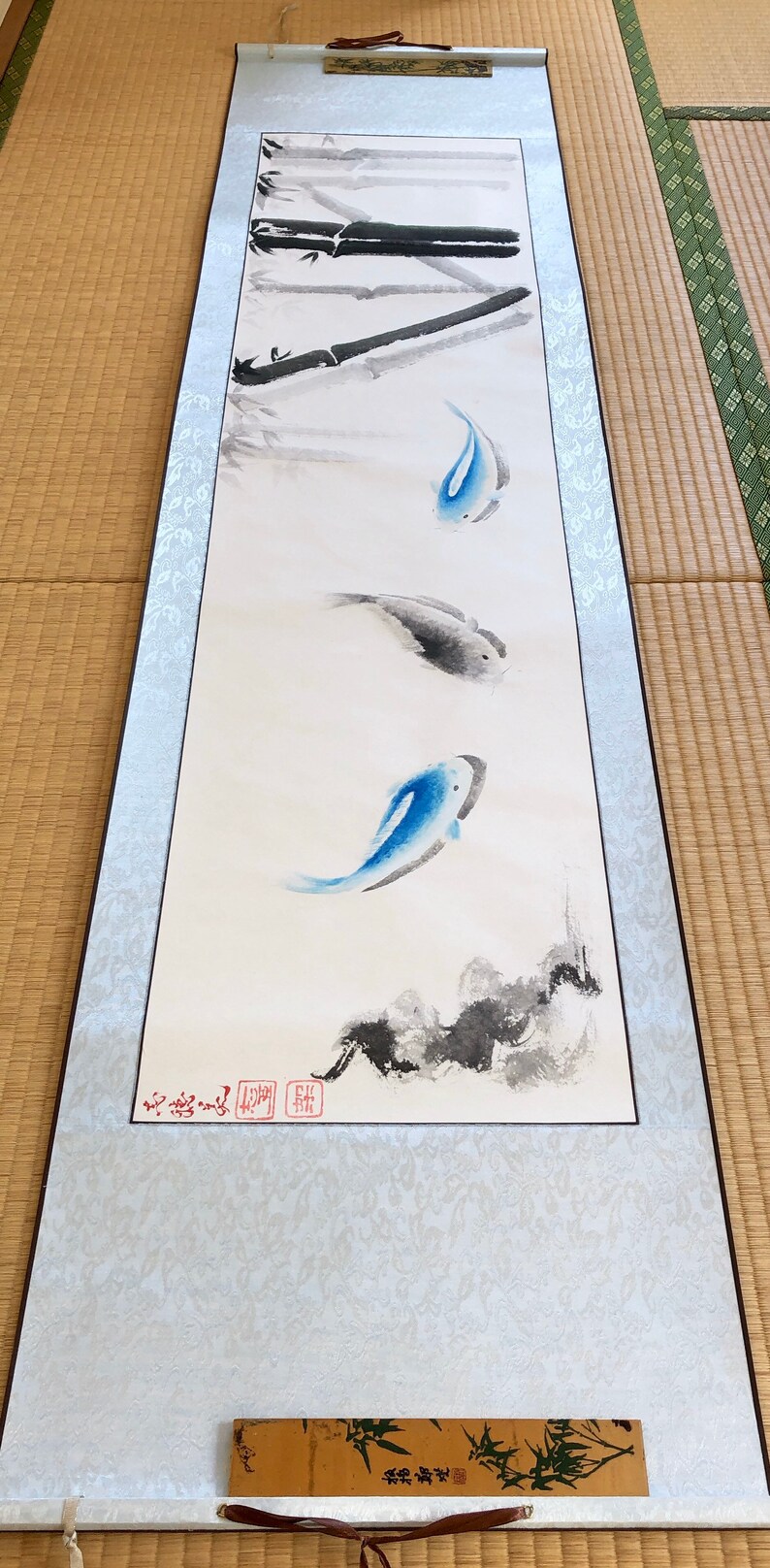 May include: A traditional Japanese scroll painting featuring three koi fish in shades of blue and gray, swimming in a minimalist style. Bamboo stalks are painted at the top. The scroll is framed with a light blue border.