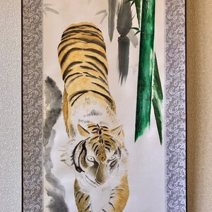 May include: A vertical painting of a tiger with orange and black stripes, walking towards the viewer. The background features bamboo stalks and leaves, with a gray floral border on the left and a signature in Japanese characters.