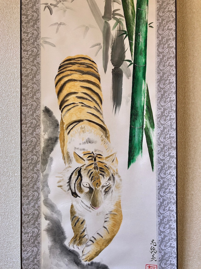 May include: A vertical painting of a tiger with orange and black stripes, walking towards the viewer. The background features bamboo stalks and leaves, with a gray floral border on the left and a signature in Japanese characters.
