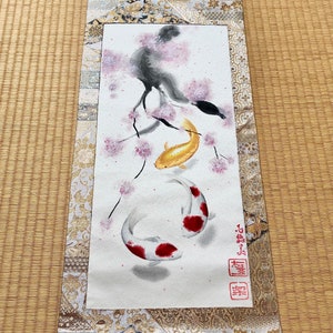 May include: Japanese scroll painting featuring two koi fish, one gold and one white with red markings, swimming beneath a cherry blossom branch. The scroll has a decorative gold and cream fabric border and wooden dowels.