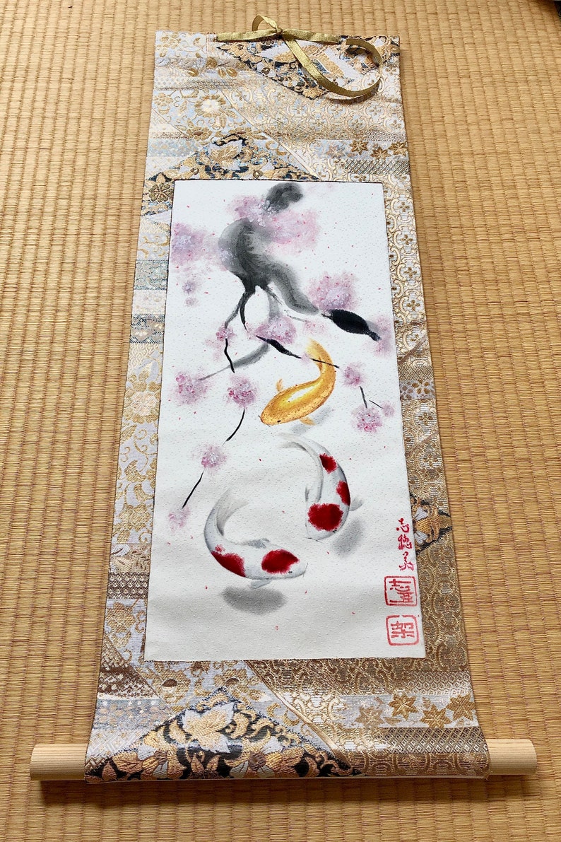 May include: Japanese scroll painting featuring two koi fish, one gold and one white with red markings, swimming beneath a cherry blossom branch. The scroll has a decorative gold and cream fabric border and wooden dowels.