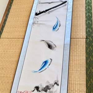 May include: A long, vertical scroll painting featuring bamboo stalks and three fish. The fish are painted in shades of blue and gray, with the bamboo in black. The scroll has a light blue border and is displayed on a tatami mat.