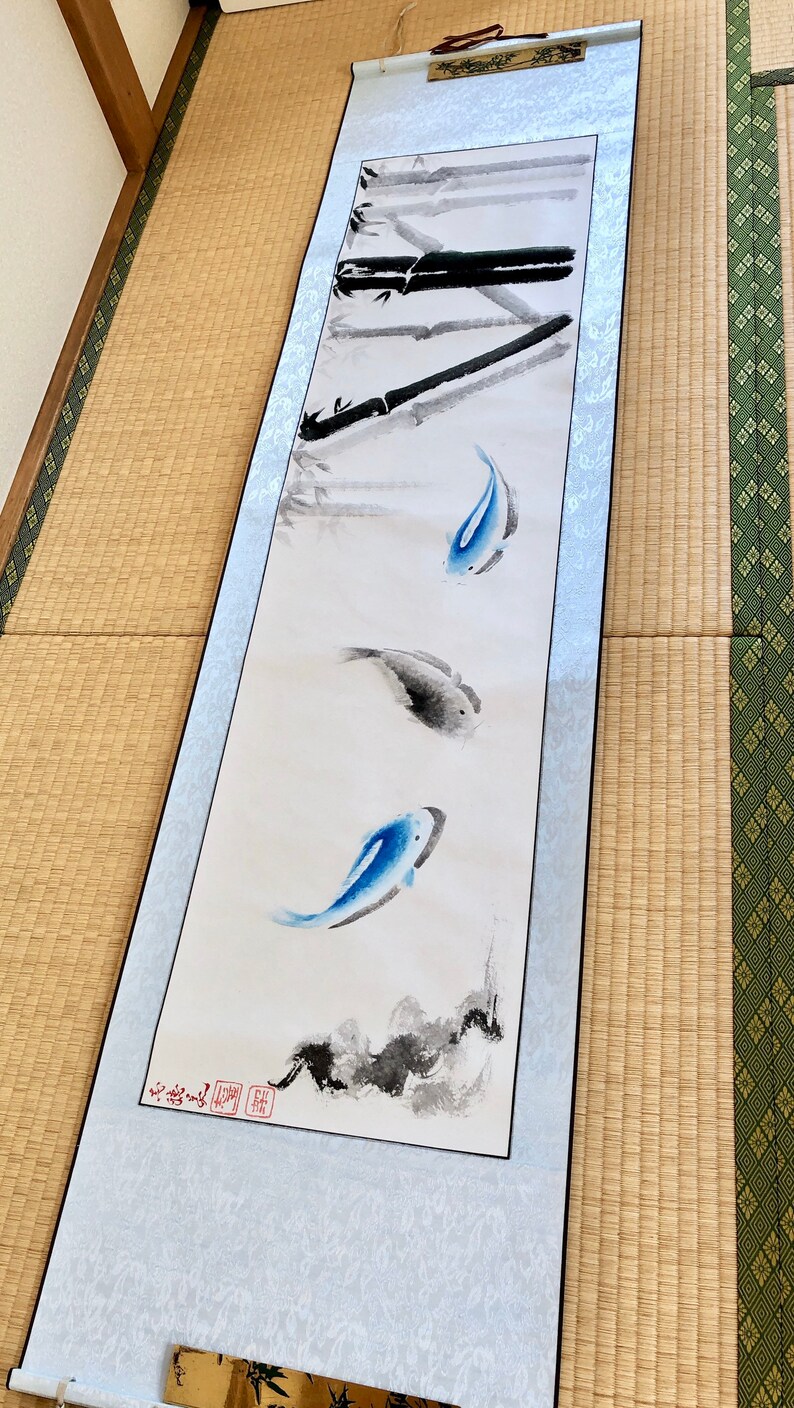 May include: A long, vertical scroll painting featuring bamboo stalks and three fish. The fish are painted in shades of blue and gray, with the bamboo in black. The scroll has a light blue border and is displayed on a tatami mat.