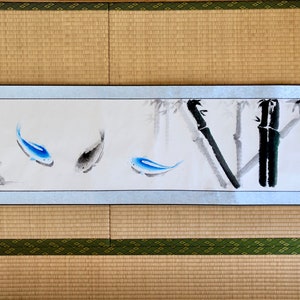 May include: A Japanese-style painting on a scroll featuring three fish in shades of blue and gray, swimming near bamboo stalks. The artwork is framed with a silver border and set against a neutral background.