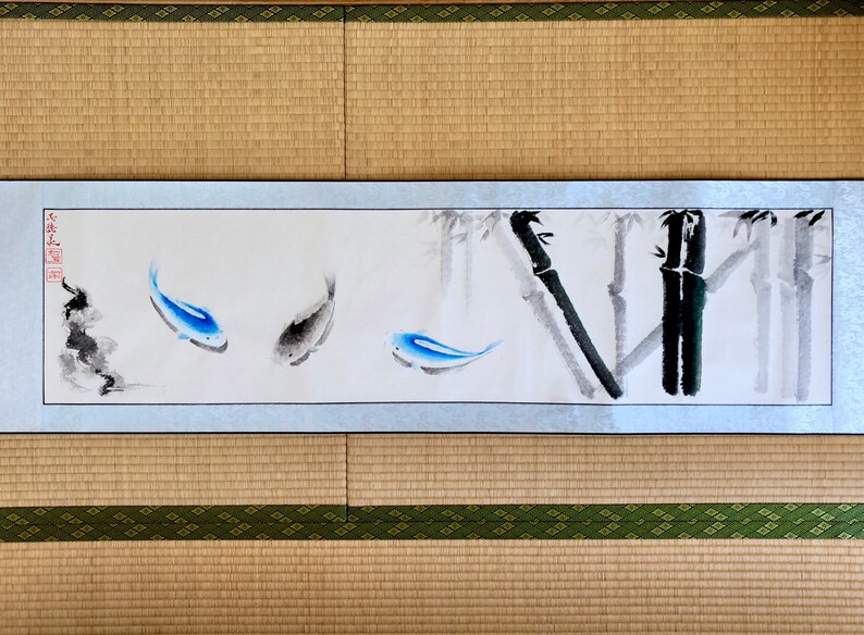 May include: A Japanese-style painting on a scroll featuring three fish in shades of blue and gray, swimming near bamboo stalks. The artwork is framed with a silver border and set against a neutral background.