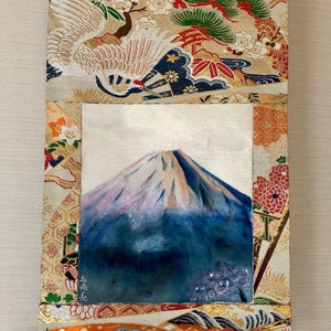 May include: A rectangular wall hanging featuring a painting of Mount Fuji with a snow-capped peak. The painting is framed by a decorative fabric with crane and floral patterns in gold, red, and green. A gold ribbon is attached for hanging.
