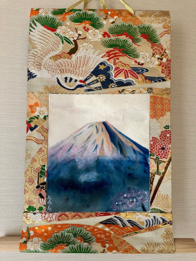 May include: A rectangular wall hanging featuring a painting of Mount Fuji with a snow-capped peak. The painting is framed by a decorative fabric with crane and floral patterns in gold, red, and green. A gold ribbon is attached for hanging.