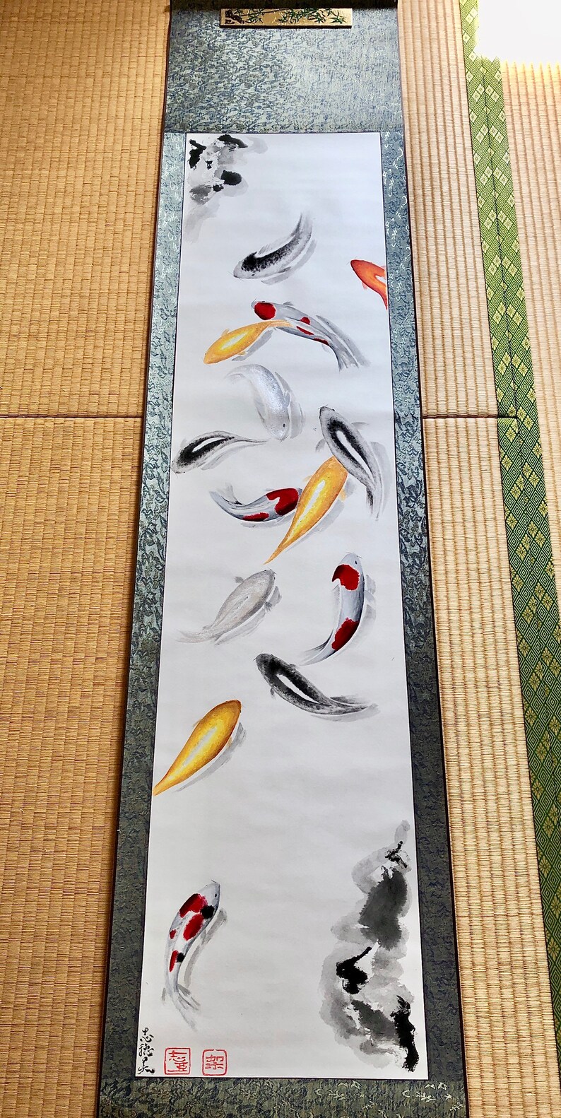 May include: A vertical scroll painting featuring koi fish in shades of black, white, red, and gold. The fish are depicted swimming in a minimalist style against a white background. The scroll has a dark border and a decorative top.