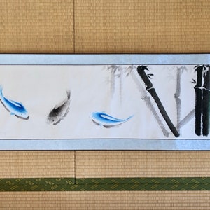 May include: Japanese-style scroll painting featuring blue and gray fish swimming near bamboo stalks. The artwork is on a light blue background with a white border. The painting is displayed on a woven mat.
