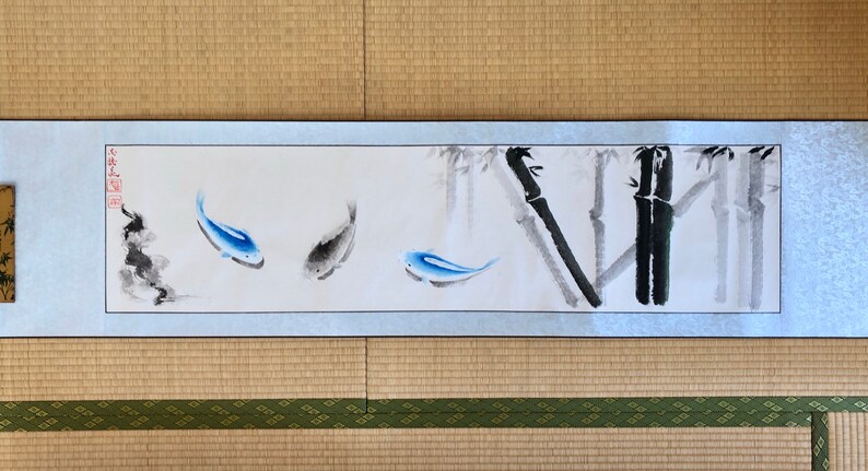 May include: Japanese-style scroll painting featuring blue and gray fish swimming near bamboo stalks. The artwork is on a light blue background with a white border. The painting is displayed on a woven mat.