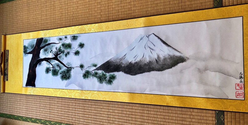 May include: Japanese-style scroll painting featuring Mount Fuji with a snow-capped peak, rendered in shades of gray and white. A tree with green foliage is on the left. The scroll has a gold border and is displayed on a woven mat.