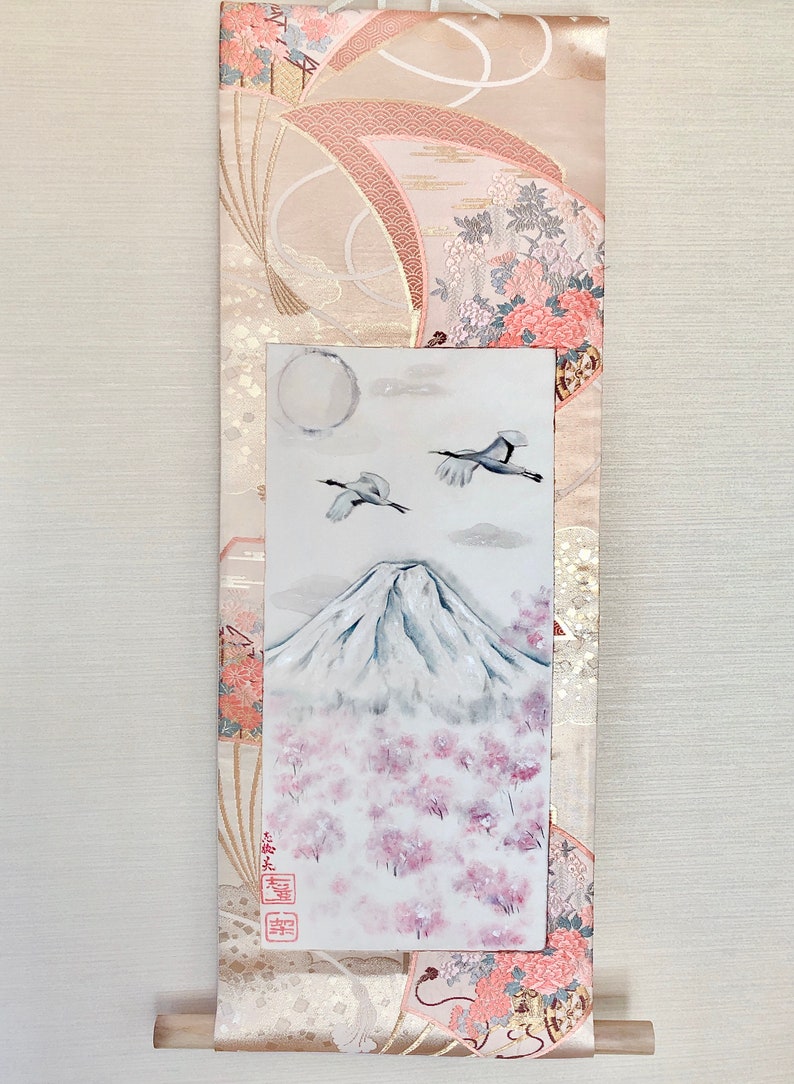 May include: Japanese wall scroll depicting Mount Fuji, cherry blossoms, and cranes. The scroll has a decorative border with floral and geometric patterns in pink, gold, and cream. The painting is framed by a patterned fabric.