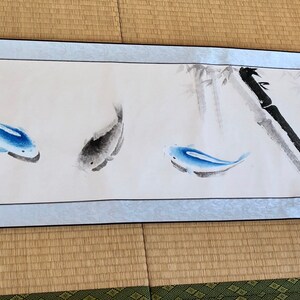 May include: A long, rectangular scroll featuring an ink wash painting. The artwork depicts three blue and gray fish swimming near bamboo stalks. The scroll has decorative wooden end pieces and is displayed on a light blue background.