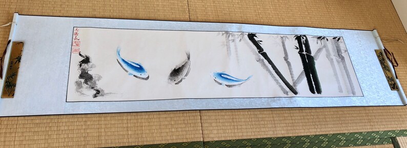May include: A long, rectangular scroll featuring an ink wash painting. The artwork depicts three blue and gray fish swimming near bamboo stalks. The scroll has decorative wooden end pieces and is displayed on a light blue background.