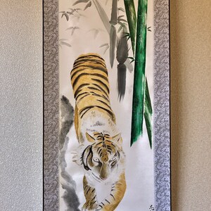 May include: Watercolor painting of a tiger with orange and black stripes, walking towards the viewer. The background features bamboo stalks and leaves in shades of green and gray. The artwork is framed with a decorative border.