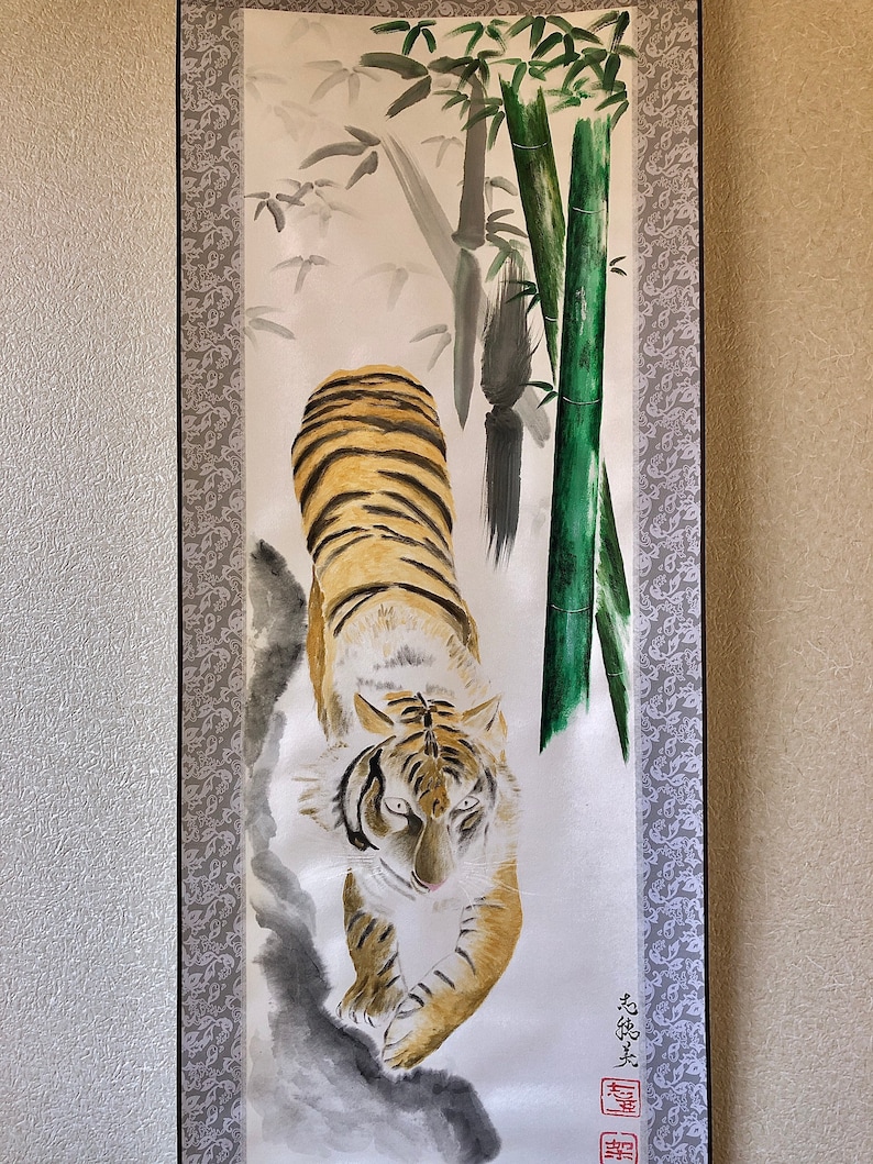 May include: Watercolor painting of a tiger with orange and black stripes, walking towards the viewer. The background features bamboo stalks and leaves in shades of green and gray. The artwork is framed with a decorative border.