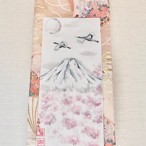May include: Japanese scroll painting depicting Mount Fuji with cherry blossoms, two cranes, and a full moon. The artwork is set against a backdrop of patterned fabric in beige, pink, and gold. The scroll has a wooden dowel at the bottom.