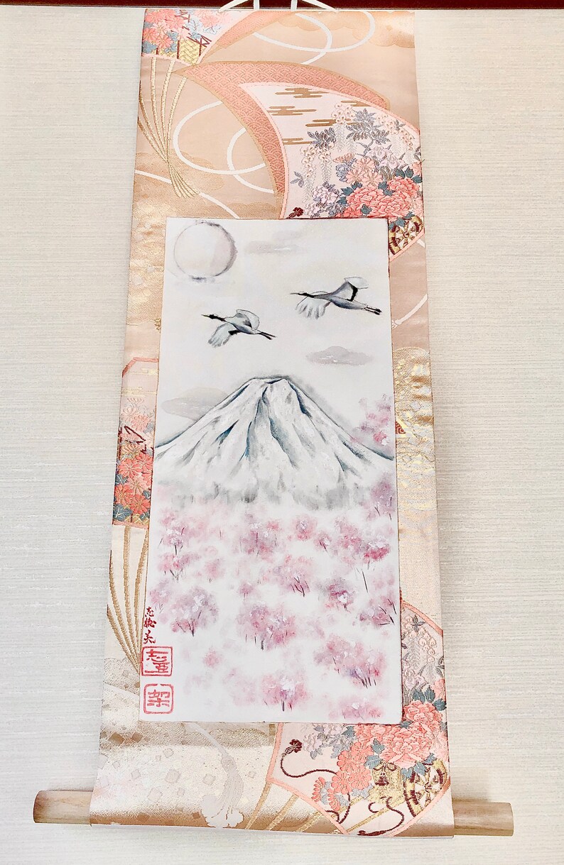 May include: Japanese scroll painting depicting Mount Fuji with cherry blossoms, two cranes, and a full moon. The artwork is set against a backdrop of patterned fabric in beige, pink, and gold. The scroll has a wooden dowel at the bottom.