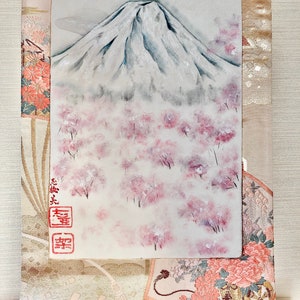 May include: A painting of Mount Fuji with a crane in flight, cherry blossoms, and Japanese characters. The artwork is set against a backdrop of a traditional patterned fabric in shades of beige, pink, and gold.