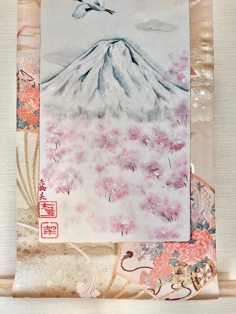May include: A painting of Mount Fuji with a crane in flight, cherry blossoms, and Japanese characters. The artwork is set against a backdrop of a traditional patterned fabric in shades of beige, pink, and gold.
