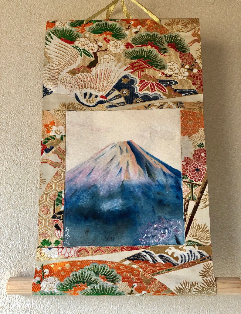 May include: A Japanese-style wall hanging featuring a painting of Mount Fuji with a snow-capped peak. The artwork is framed by ornate fabric panels with gold, orange, and cream colors, and traditional Japanese motifs.