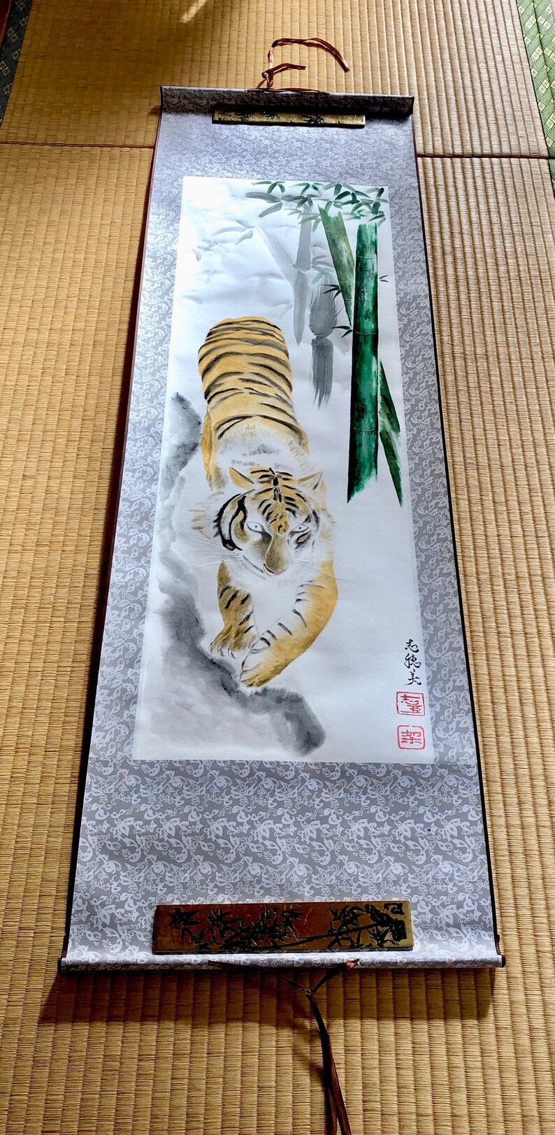 May include: A traditional Japanese scroll painting featuring a tiger with orange and black stripes, walking towards the viewer. Bamboo stalks and foliage are in the background. The scroll has a gray patterned border and wooden accents.