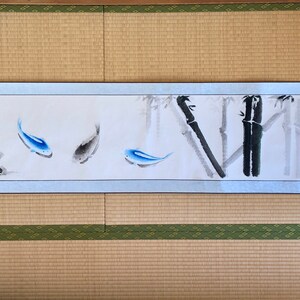May include: A Japanese-style scroll painting featuring three fish in shades of blue and gray, with bamboo stalks in the background. The artwork is framed by a light blue border and includes Japanese characters. The scroll is displayed on a traditional tatami mat.