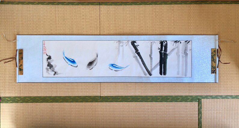 May include: A Japanese-style scroll painting featuring three fish in shades of blue and gray, with bamboo stalks in the background. The artwork is framed by a light blue border and includes Japanese characters. The scroll is displayed on a traditional tatami mat.