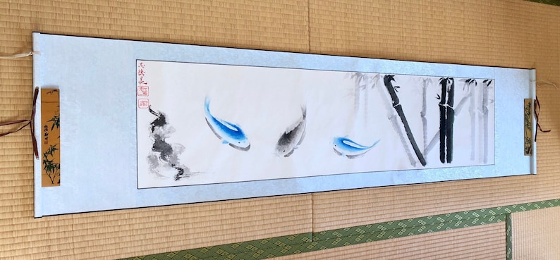 May include: A long, rectangular scroll featuring a watercolor painting of three fish and bamboo stalks. The fish are blue and gray, and the bamboo is black and gray. The scroll has a light blue border and is displayed on a woven mat.