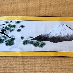 May include: A Japanese scroll painting featuring Mount Fuji with a snow-capped peak, a dark tree, and green foliage. The artwork is framed by a gold border and has decorative end pieces. The scroll is displayed on a tatami mat.