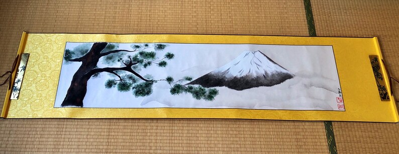 May include: A Japanese scroll painting featuring Mount Fuji with a snow-capped peak, a dark tree, and green foliage. The artwork is framed by a gold border and has decorative end pieces. The scroll is displayed on a tatami mat.