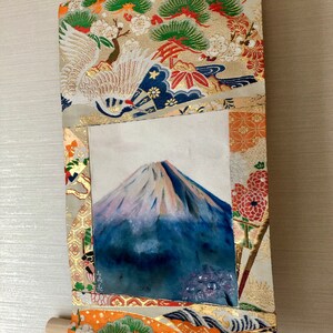 May include: Japanese-style wall scroll featuring a painting of Mount Fuji with a snow-capped peak. The scroll is framed with ornate fabric in gold, orange, and cream colors, with crane and pine tree motifs. A wooden dowel is at the bottom, and a gold ribbon hangs from the top.