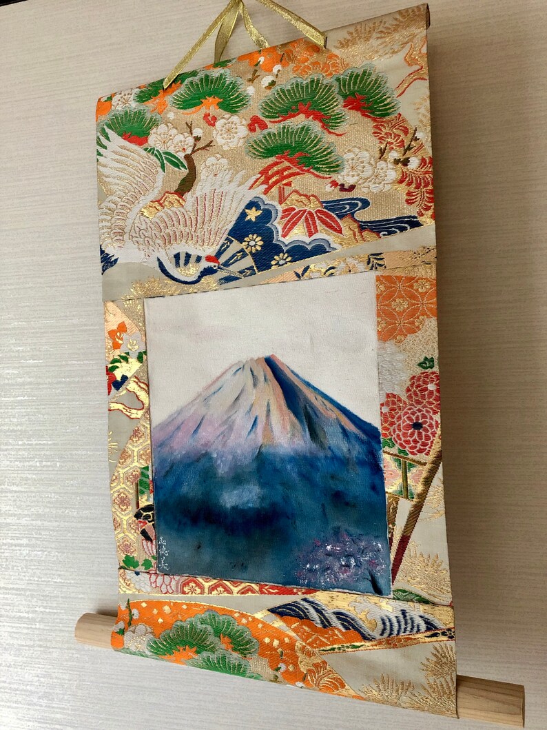 May include: Japanese-style wall scroll featuring a painting of Mount Fuji with a snow-capped peak. The scroll is framed with ornate fabric in gold, orange, and cream colors, with crane and pine tree motifs. A wooden dowel is at the bottom, and a gold ribbon hangs from the top.