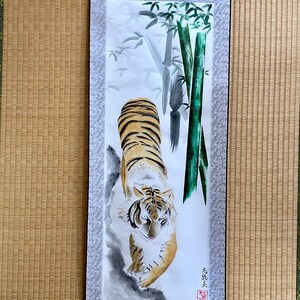 May include: A vertical scroll painting featuring a tiger with orange and black stripes, walking towards the viewer. Bamboo stalks are on the right. The scroll has a gray patterned border and a brown hanging cord.