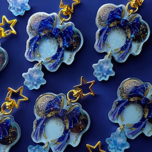May include: A set of blue and gold charm keychains featuring a blue bow with gold accents and a star charm. The charms are on a clear background with a sparkly effect.